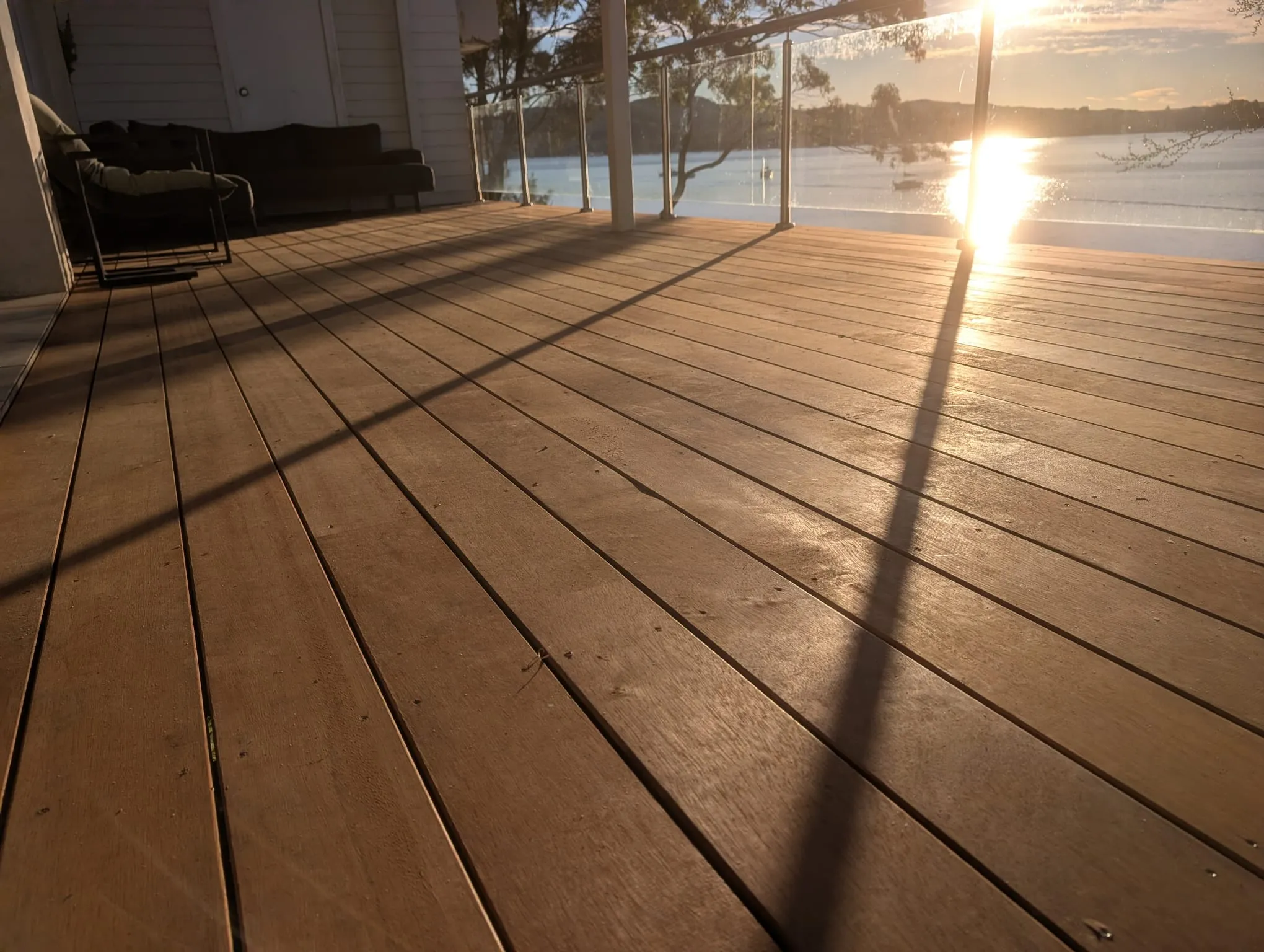 A renovated timber deck with a glass balustrade, lit by the setting sun over the water